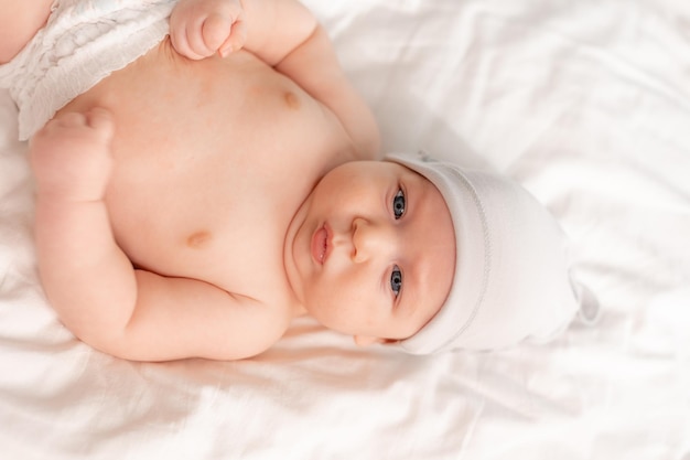 Портрет младенца с голубыми глазами в подгузнике и белой шапочке, лежащего на спине на белом постельном белье. место для текста. Фото высокого качества