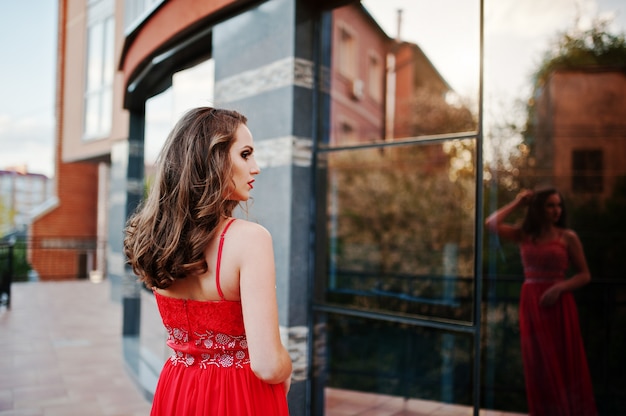 Portrait back of fashionable girl at red evening dress posed background mirror window of modern building