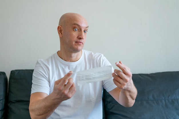 Portrait of a bald man sitting at home on the sofa holding a medical mask in his hands