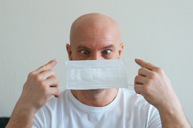 Portrait of a bald man sitting at home on the sofa holding a medical mask in his hands