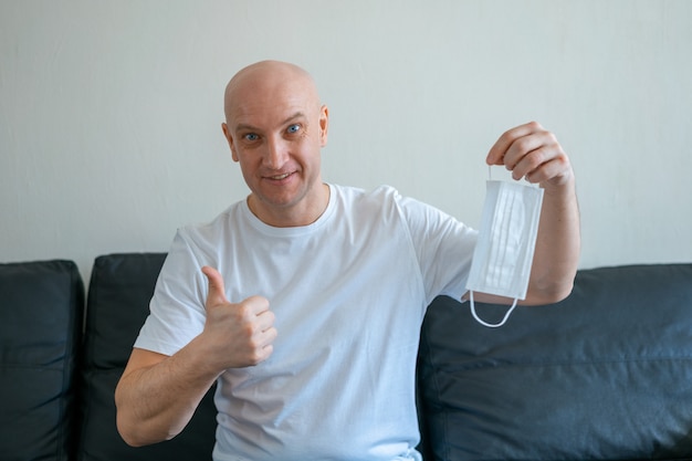 Photo portrait of a bald man sitting at home on the sofa holding a medical mask in his hands