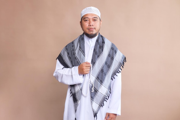 Portrait of bearded Asian muslim man in white robe and cap holding prayer beads