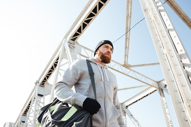 Foto ritratto di un atleta barbuto con borsa sportiva che cammina lungo il ponte urbano