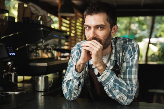 Портрет бородатого парня-бариста в фартуке, улыбающегося во время работы в уличном кафе или кофейне на открытом воздухе