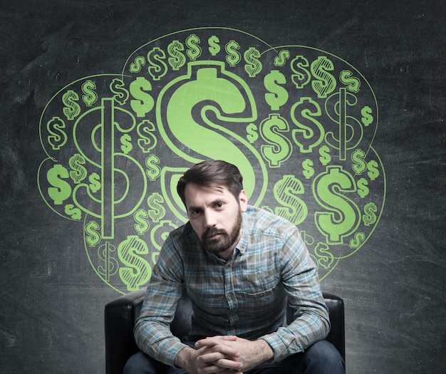 Portrait of a bearded businessman wearing a checkered shirt and standing with an open mouth near a blackboard with dollar signs drawn on it.