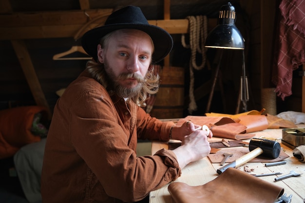 Portrait of bearded carpenter in hat looking at camera while sitting at his workplace and working with tools in workshop