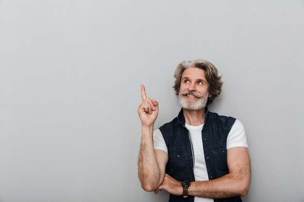 Portrait of a bearded cheery smiling senior man with grey hair isolated over grey wall background pointing aside.