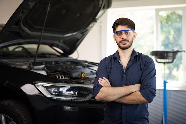 Portrait bearded handsome car mechanic is posing in a car service he wears a jeans shirt and safety