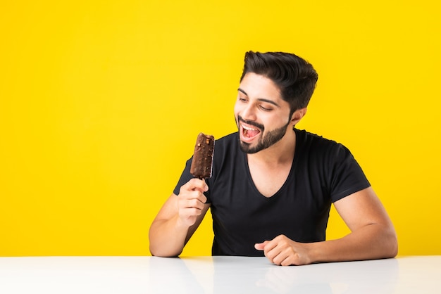 Portrait of a bearded handsome indian young manãâ eating ice cream in cone or popsicle while sitting at table against yellow studio background