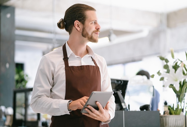 Photo portrait of bearded hipster barista using digital tablet and looking away while standing at trendy coffee shop happy barista man are taking orders from online customers startup small business owner