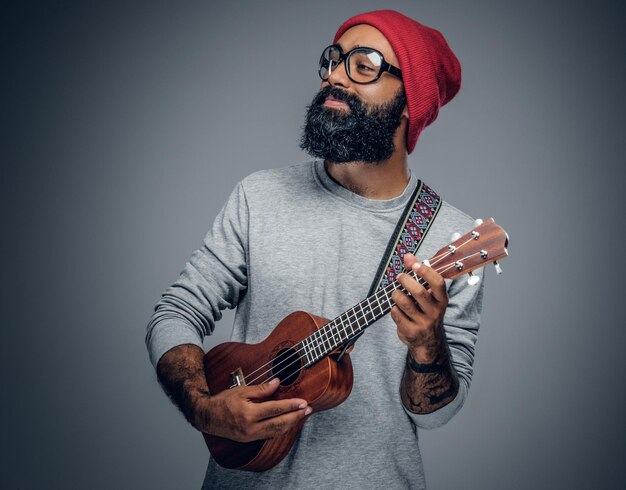 Portrait of a bearded hipster male in red hat playing on ukulele. Isolated on grey background.