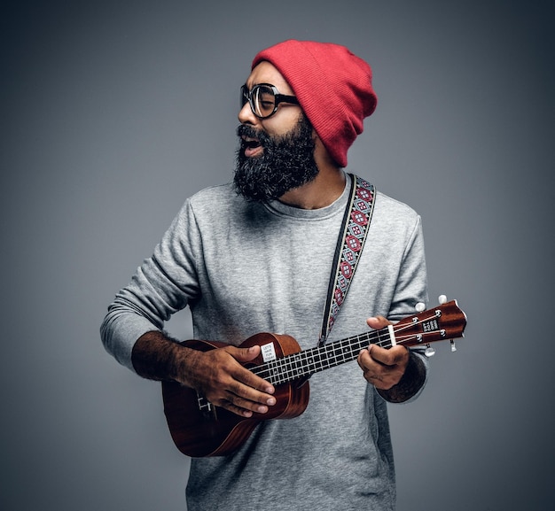 Portrait of a bearded hipster male in red hat playing on ukulele. Isolated on grey background.