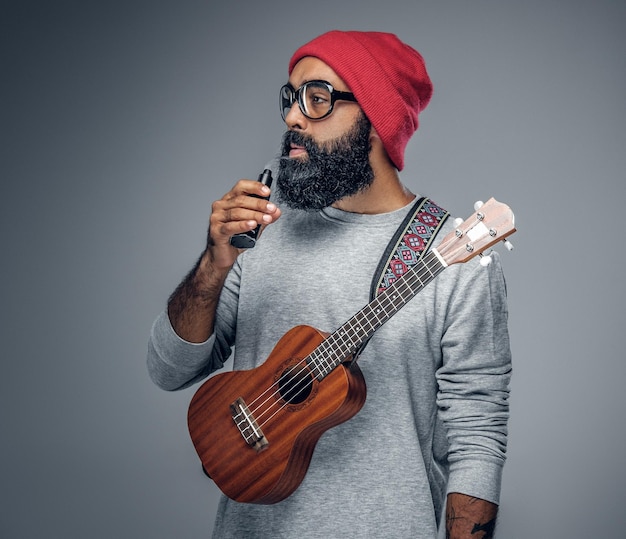 Portrait of a bearded hipster male in red hat playing on ukulele. Isolated on grey background.