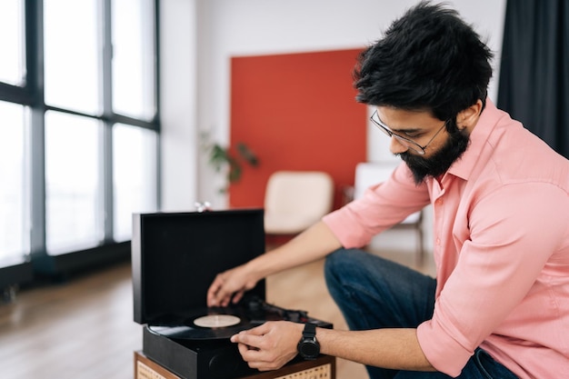 Portrait of bearded Indian hipster in glasses adjusting head of old record player and vinyl discs sitting on floor Music lover man listening to classical musi