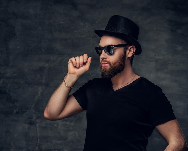 Portrait of bearded male in a black t shirt, cylinder hat and sunglasses over grey background.