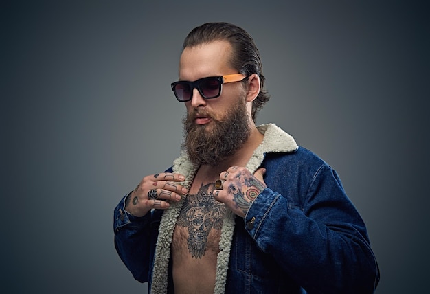 Portrait of bearded male in sunglasses with tattoo on a chest wearing a denim jacket.