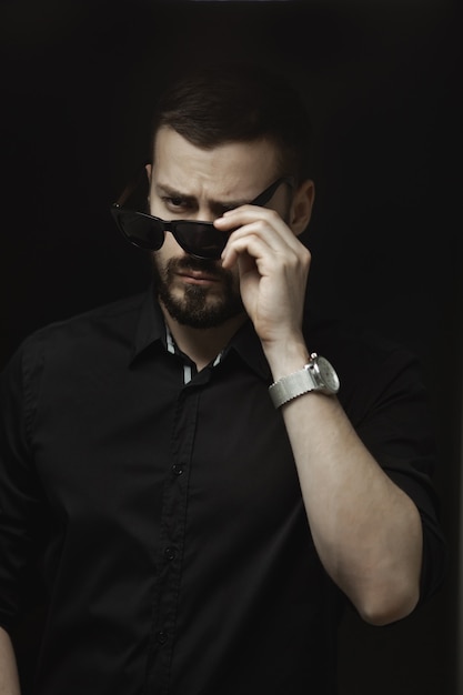 Портрет бородатого мужчины в черной рубашке и солнечных очках с напряженным взглядом, стоящего в помещении