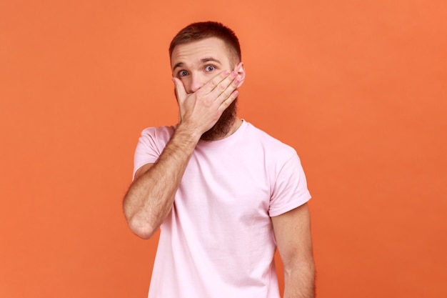 Portrait of bearded man covering mouth with hand to keep silent afraid to say secret looking with intimidated expression wearing pink Tshirt Indoor studio shot isolated on orange background