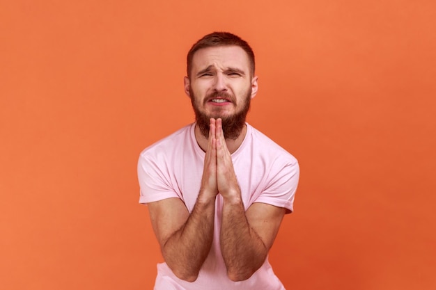 Portrait of bearded man folding hands in prayer closing eyes and talking to god asking for help expressing gratitude wearing pink Tshirt Indoor studio shot isolated on orange background