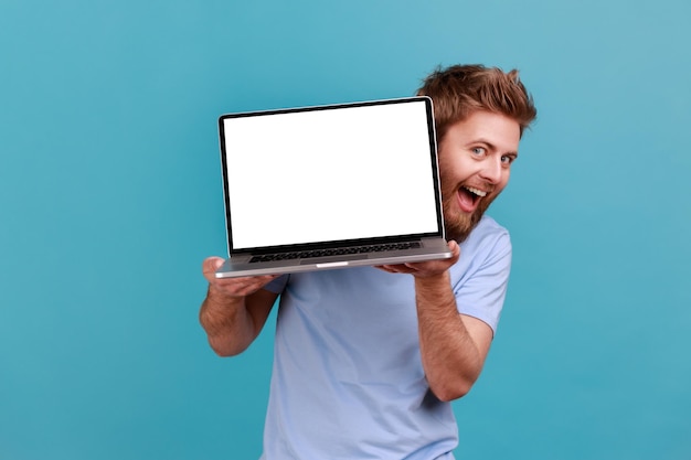 Portrait of bearded man holding laptop with blank white screen and looking at camera with excited facial expression and toothy smile. Indoor studio shot isolated on blue background.