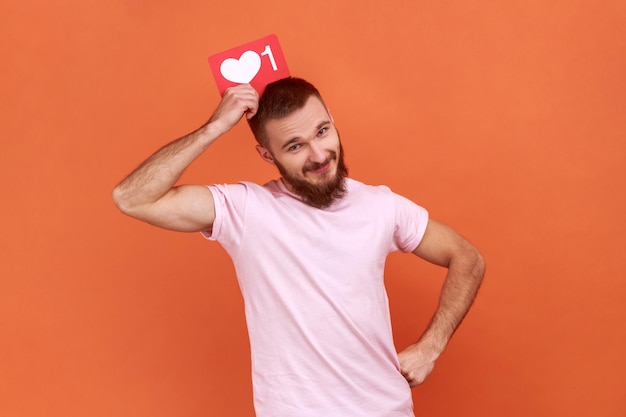 Ritratto di uomo barbuto che tiene come icona sopra la testa guardando la fotocamera con espressione facciale soddisfatta social media che indossa una maglietta rosa indoor studio girato isolato su sfondo arancione