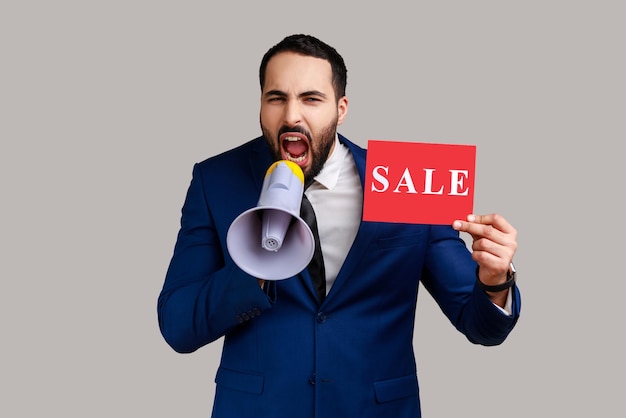 Portrait of bearded man paying attention announcing big sale yelling at loudspeaker, black Friday shopping, wearing official style suit. Indoor studio shot isolated on gray background.