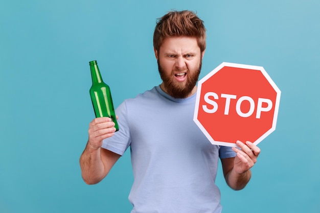 Portrait of bearded man showing alcoholic beverage beer bottle\
and stop sign warning and worrying looking at camera with angry\
expression indoor studio shot isolated on blue background