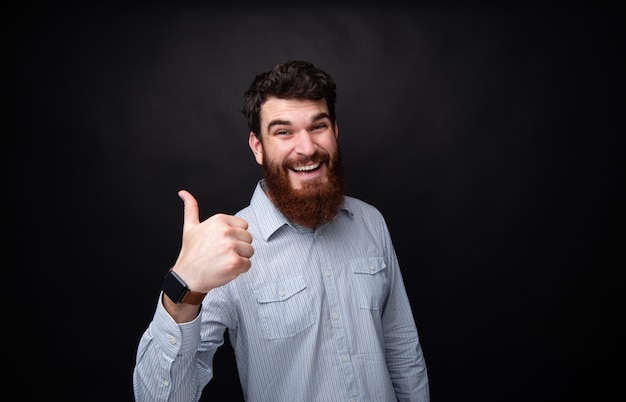 Photo portrait of bearded man showing thumb up with toothy smile over isolated dark background