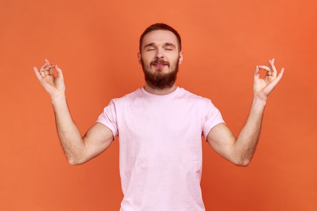 Портрет бородатого мужчины, стоящего с поднятыми руками и занимающегося йогой, медитацией, жестом мудры, в розовой футболке. Крытая студия снята на оранжевом фоне.