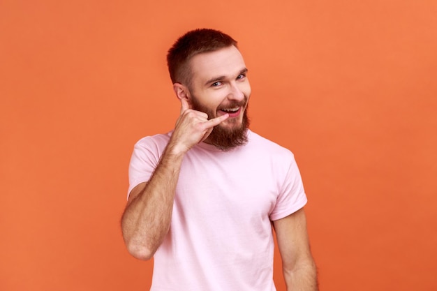 Portrait of bearded man standing with telephone hand gesture and smiling to camera flirting offering to contact by phone wearing pink Tshirt Indoor studio shot isolated on orange background