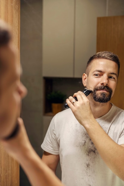 Portrait of a bearded man styling his beard with shaver in bathroom