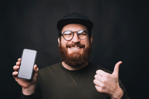 portrait of a bearded man wearing glasses showing thumb up and his phone.