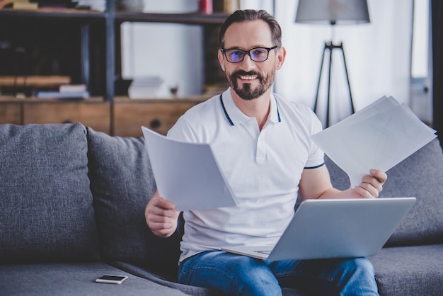 Portrait of bearded man wearing glasses working on laptop holding documents at home and looking at the camera