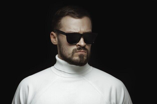 Portrait of a bearded man in a white sweater and sunglasses with an intense look standing indoors