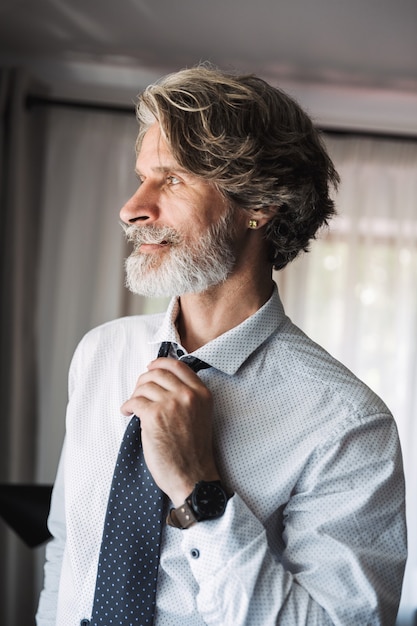 Portrait of a bearded mature handsome grey-haired businessman posing indoors at home near window dressed in formal clothes correct his tie.