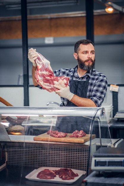 Portrait of a bearded meat man dressed in a fleece shirt holds fresh cut meat in a market.