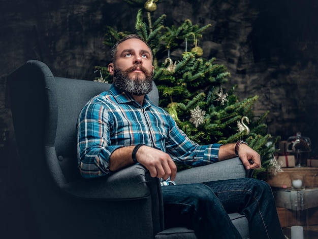 Portrait of bearded middle age male dressed in plaid flannel shirt and jeans sits on a chair over Christmas decorated background.