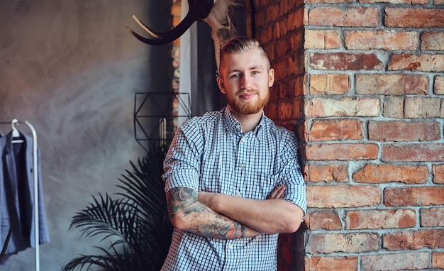 Portrait of a bearded modern male with tattoos on his arms posing near the window in a room with loft interior.