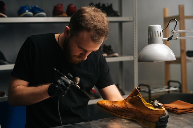 Portrait of bearded shoemaker wearing black gloves spraying paint of light brown leather shoes closeup