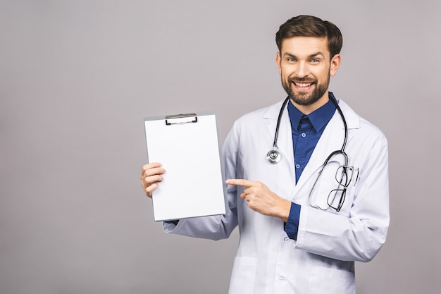 Portrait of bearded smiling intern, who is holding the clipboard with empty paper. Doctor is wearing white uniform, stands over isolated grey background, showing blank.