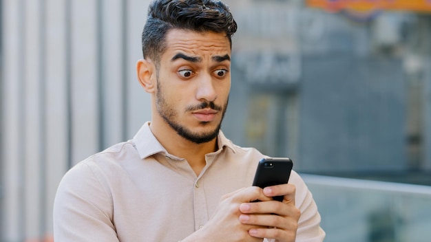 수염을 기른 젊은 미남 사업가가 얼굴에 놀란 표정으로 휴대전화를 쳐다보며 도시에 서서 메시지 충격 공포증을 받는 나쁜 소식을 읽는다.