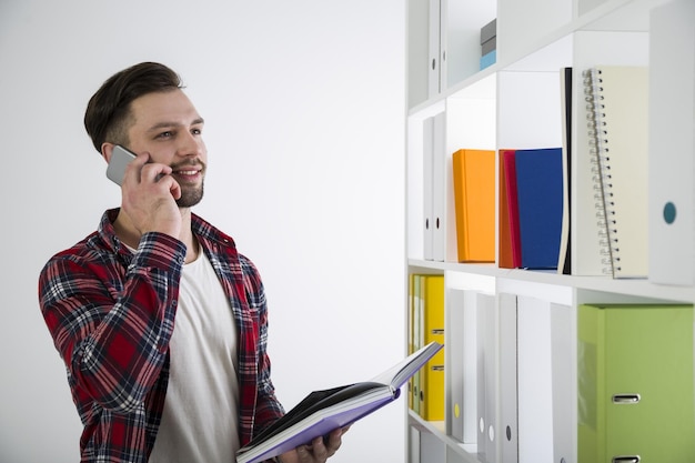 Portrait of a bearded young man in a library. He is wearing a checkered shirt and talking on his smartphone