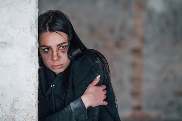 Portrait of beaten young woman with bruise under eye that standing and leaning on the wall indoors in abandoned building