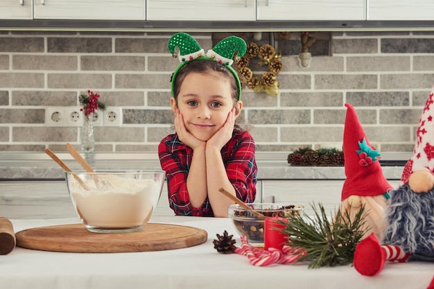 Ritratto di una bellissima adorabile bambina europea con un cerchio da elfo sulla testa sorride graziosamente guardando la telecamera seduta a un tavolo della cucina con ingredienti per cucinare una torta di natale