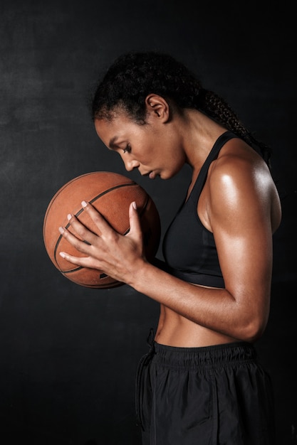 Ritratto di bella donna afroamericana in abbigliamento sportivo che tiene pallacanestro isolata sul nero