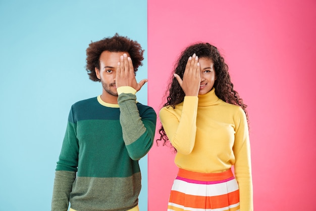 Photo portrait of beautiful african american young couple covered halves of their faces by hands over colorful background