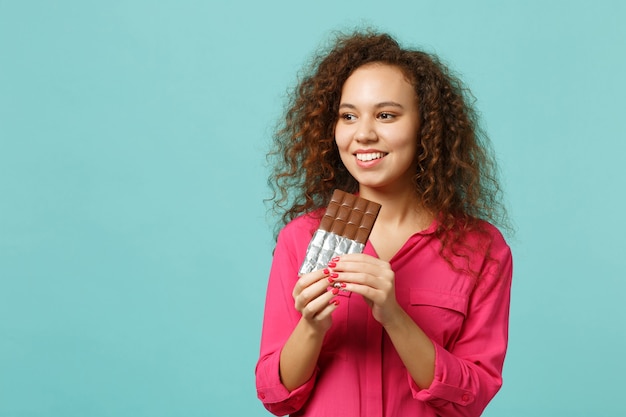 Portrait of beautiful african girl in casual clothes holding in hand chocolate bar isolated on blue turquoise wall background in studio. People sincere emotions, lifestyle concept. Mock up copy space.