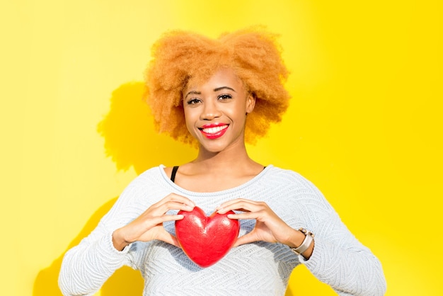 Photo portrait of a beautiful african woman holding red heart on the yellow background. valentine's day concept