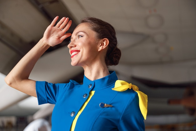 旅客機の前に立って、目をそらしながら笑っている明るい青色の制服を着た美しいエアスチュワーデスの肖像画。職業コンセプト