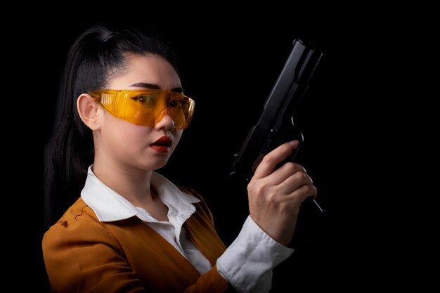 黒い壁にピストル銃を持っている黄色のスーツ片手で身に着けている美しい海の女性の肖像画、カメラを見て拳銃で若いセクシーな女の子の長い髪、きれいな女性がピストルで立っています。